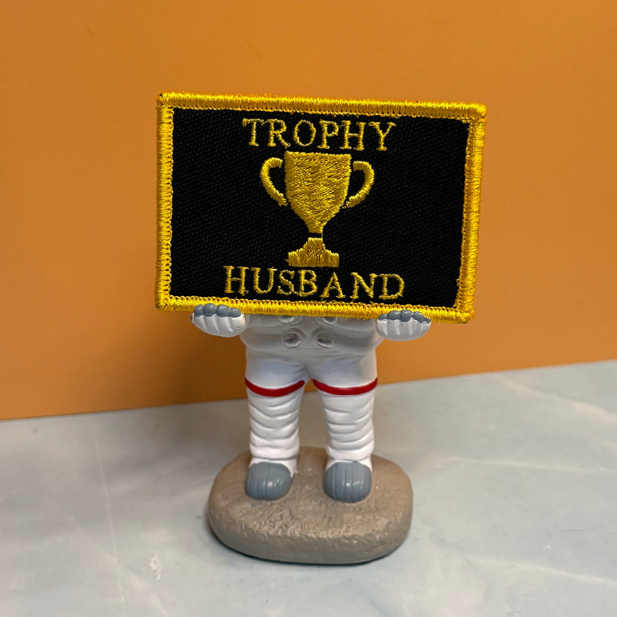 An embroidered patch with a black background, gold border, and gold trophy graphic with the words 'TROPHY HUSBAND' in gold, held by a small white astronaut figurine on a gray base against an orange and gray background.