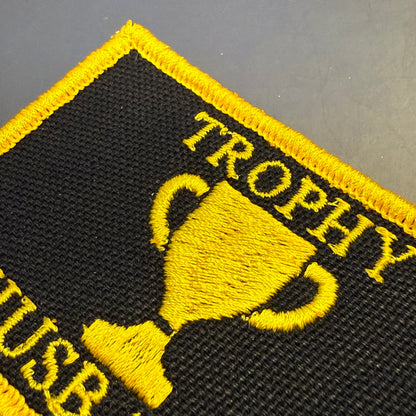 Close-up of a black embroidered patch with a yellow border, yellow trophy graphic, and partial yellow text reading 'TROPHY' and 'USB' on a gray surface.