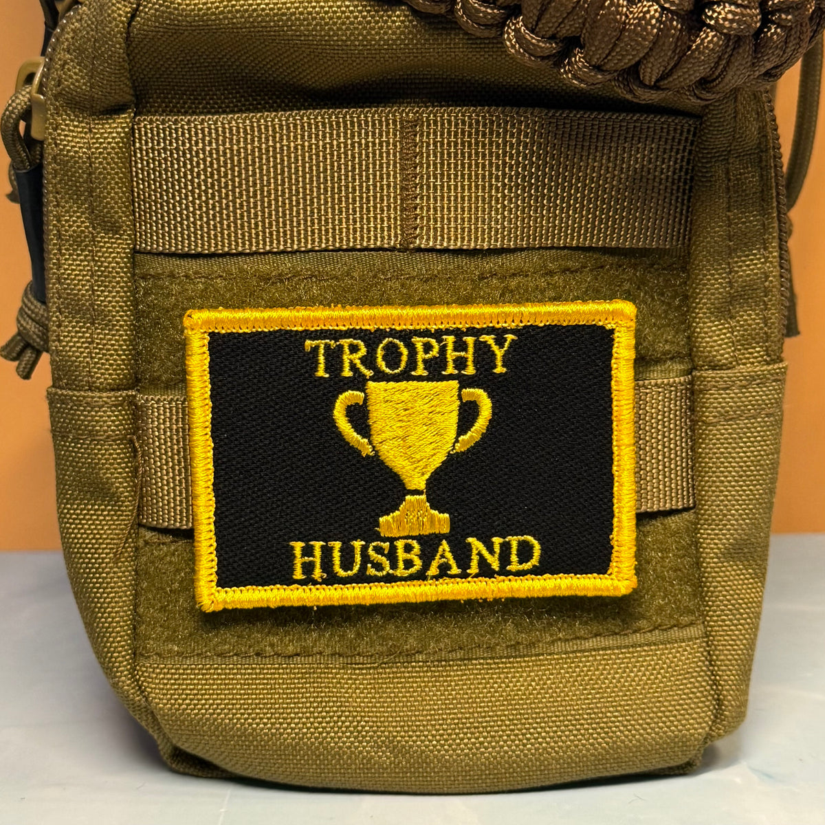 A close-up of a tactical pouch in olive drab green nylon, featuring a rectangular black patch with a yellow border and the embroidered text 'TROPHY HUSBAND' surrounding a yellow trophy graphic.