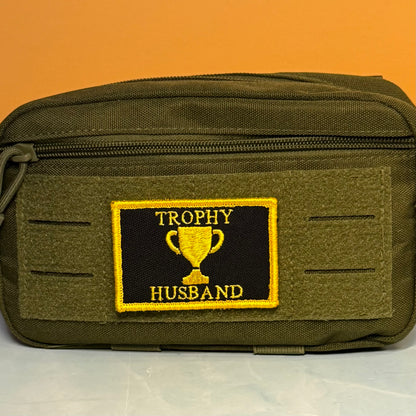 An olive green tactical pouch with a front zipper and a rectangular embroidered patch reading 'TROPHY HUSBAND' with a gold trophy emblem on a black background.