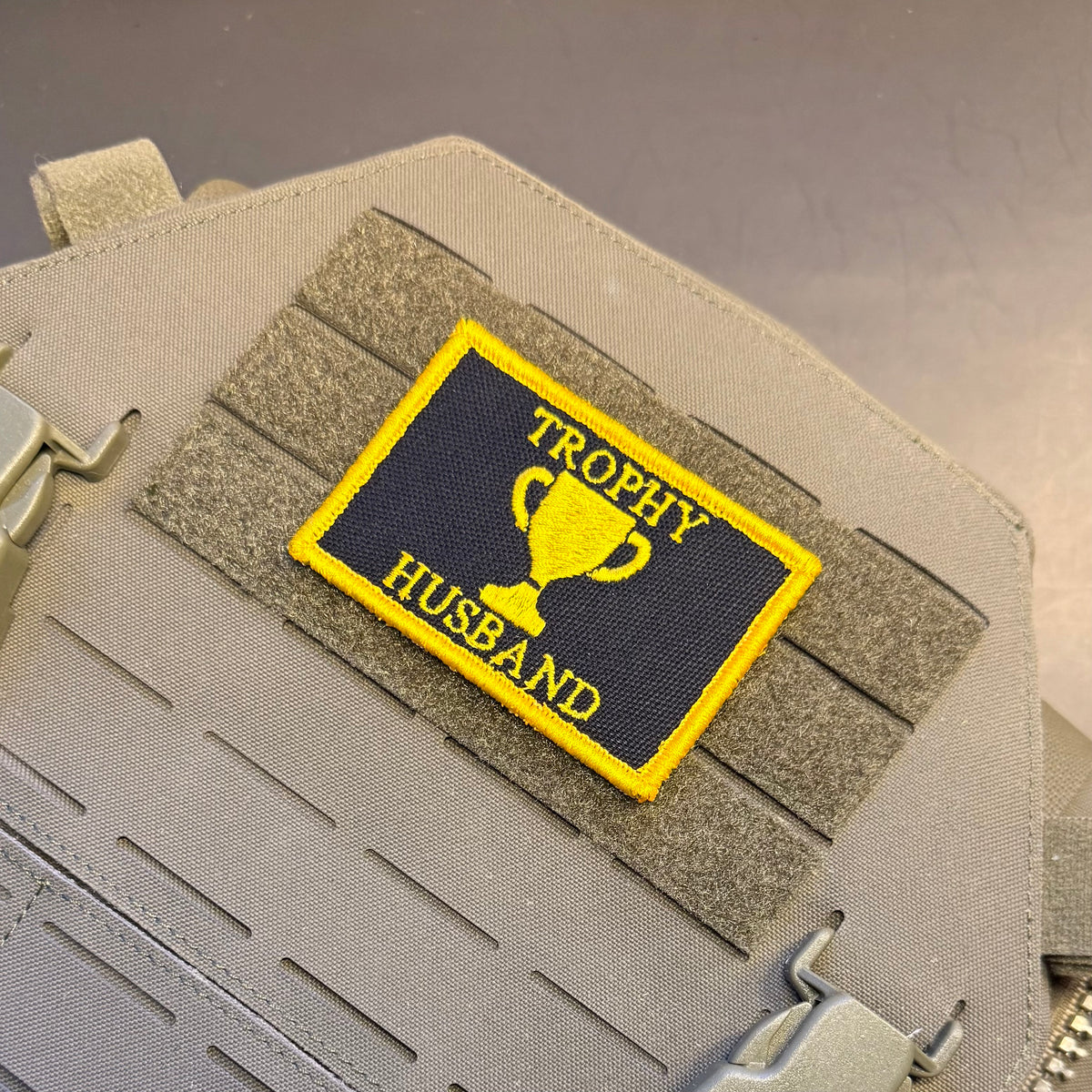 A rectangular embroidered patch with a yellow border and yellow text reading 'TROPHY HUSBAND' with a trophy graphic, attached to a tan tactical vest or bag with hook-and-loop fastening.