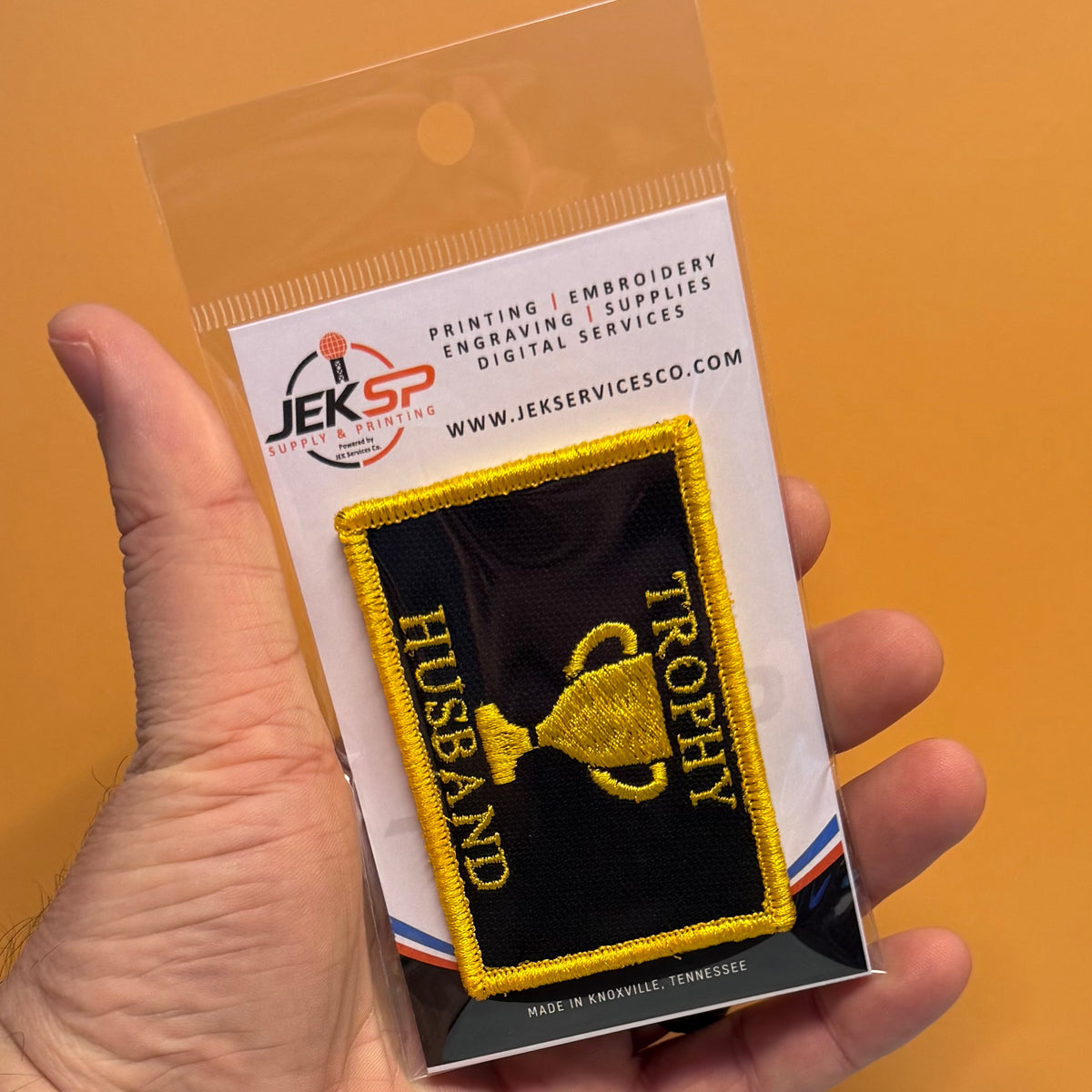 A hand holds a clear plastic blister pack containing an embroidered patch. The patch is black with yellow border and yellow text reading 'HUSBAND' and 'TROPHY' around a yellow trophy graphic.