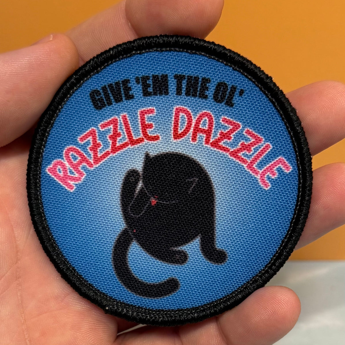 A round embroidered patch held in a hand, featuring a blue background with the text 'GIVE 'EM THE OL' RAZZLE DAZZLE' in black and red, and a black cat illustration.