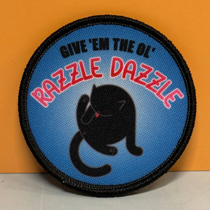 A circular embroidered patch with a black stitched border, a blue background, a black cat silhouette, and red-and-white outlined text reading “RAZZLE DAZZLE,” with smaller black text above reading “GIVE 'EM THE OL”.