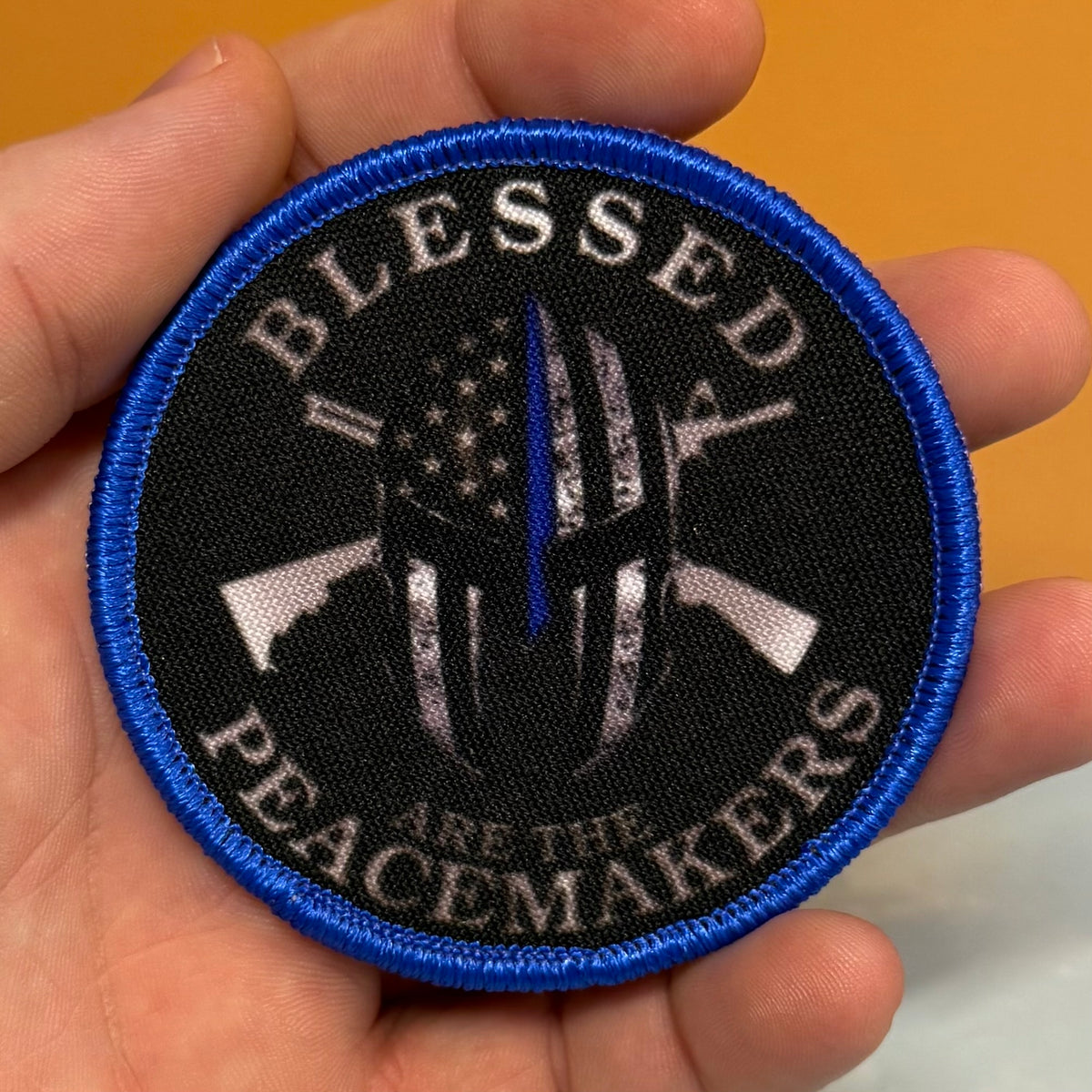 A circular embroidered patch held in a hand, featuring a black background with a blue border, white text reading 'BLESSED PEACEMAKERS', and a central peace-themed graphic with crossed rifles and a distressed American flag with a single blue stripe.