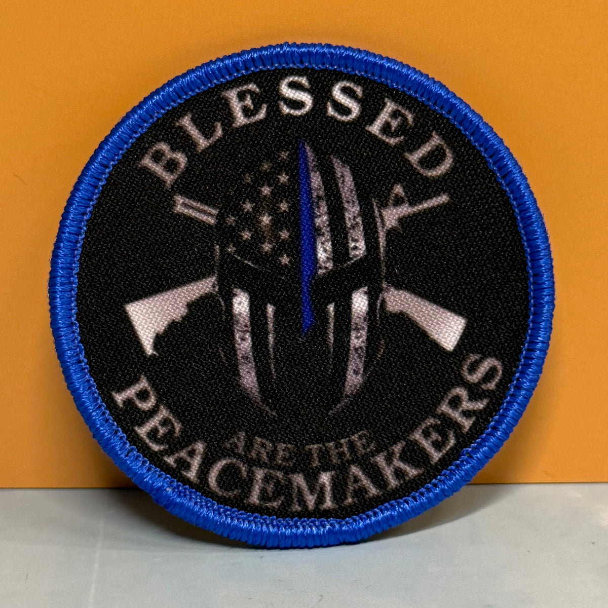 A circular embroidered patch with a blue border, black background, and a central Spartan helmet design with a vertical blue line resembling a flagpole. The patch reads “BLESSED PEACEMAKERS” in white text and is photographed against an orange background.
