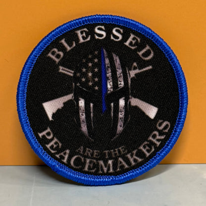 A circular embroidered patch with a blue border, black background, and a central Spartan helmet design with a vertical blue line resembling a flagpole. The patch reads “BLESSED PEACEMAKERS” in white text and is photographed against an orange background.