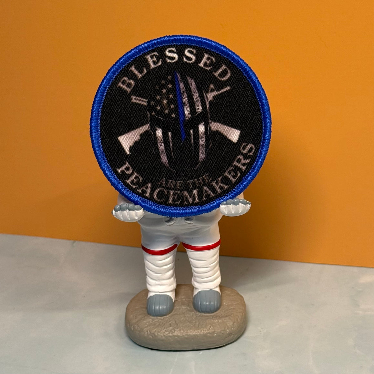 A circular embroidered patch with a blue border and the text 'BLESSED PEACEMAKERS' around a black central design, displayed on top of a small astronaut figurine against an orange and gray background.