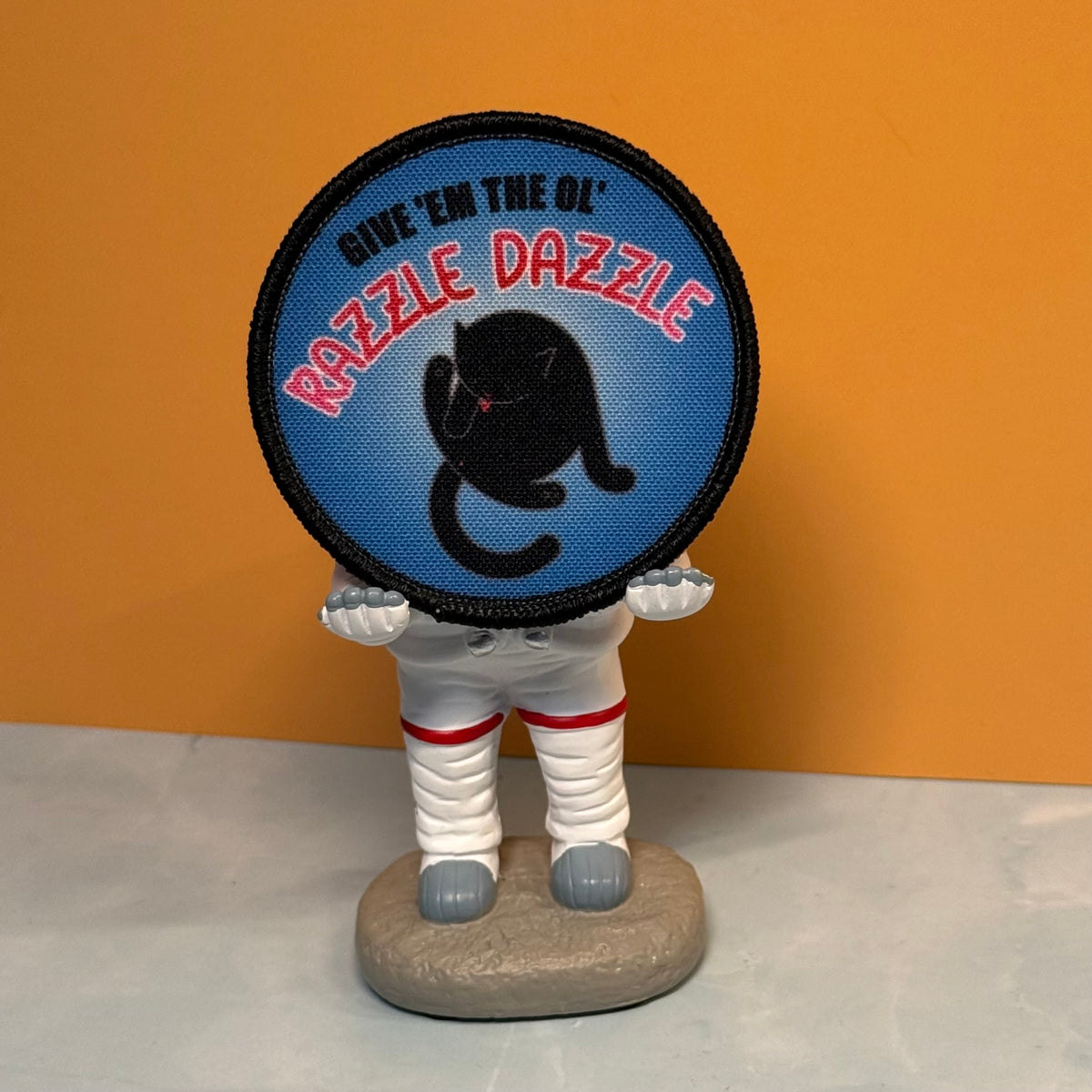 A round embroidered patch with a black cat silhouette and the text “GIVE ’EM THE OL’ RAZZLE DAZZLE” on a blue background, held by a small astronaut figurine against an orange and gray backdrop.