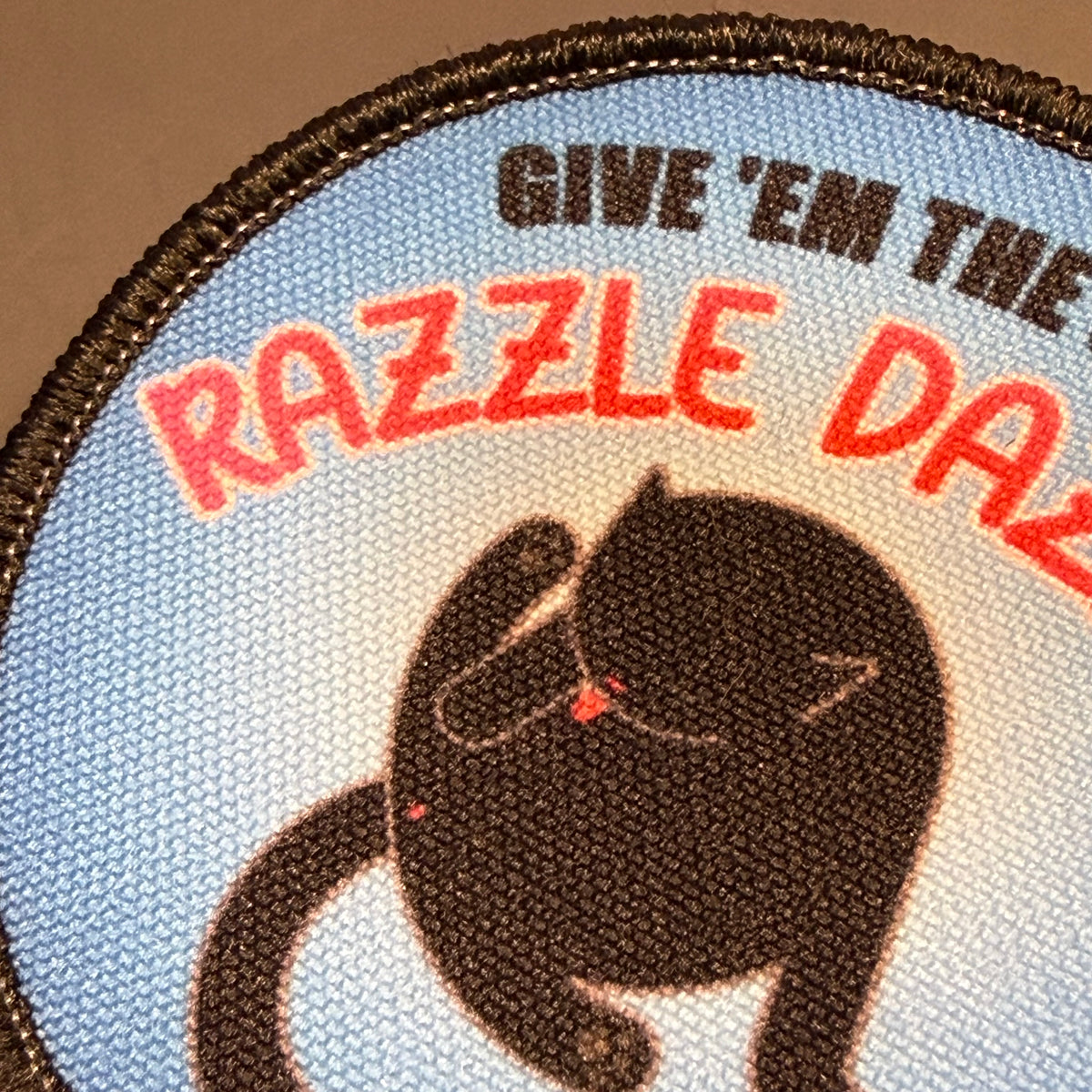 A close-up of a circular embroidered patch featuring a black cat on a light blue background with red and black text reading “GIVE ’EM THE RAZZLE DAZZLE.”.