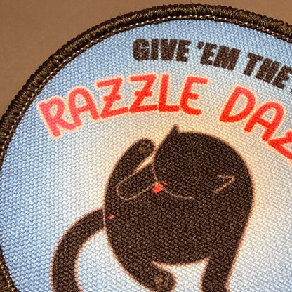 A close-up of a circular embroidered patch featuring a black cat on a light blue background with red and black text reading “GIVE ’EM THE RAZZLE DAZZLE.”.