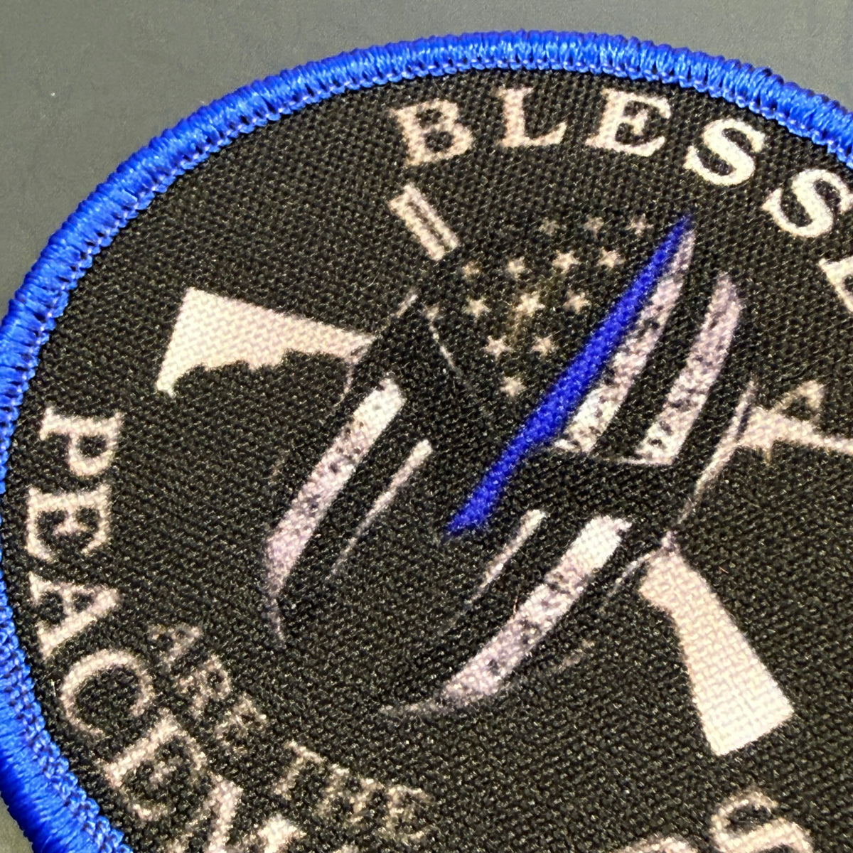 A close-up of a circular embroidered patch with a blue stitched border, black background, and a distressed American flag design with a single blue stripe. Partial text reads 'BLESSED' at the top and 'PEACE' at the side.