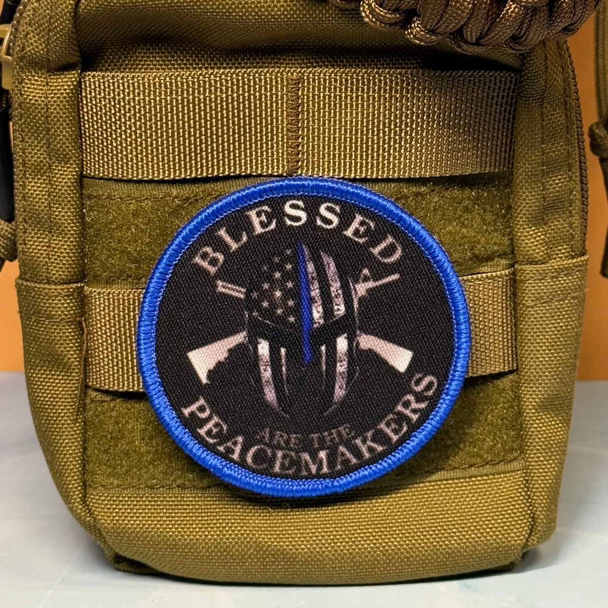 A circular embroidered tactical patch with the text 'BLESSED PEACEMAKERS' and a black, white, and blue distressed American flag helmet design, shown attached to an olive drab MOLLE pouch.