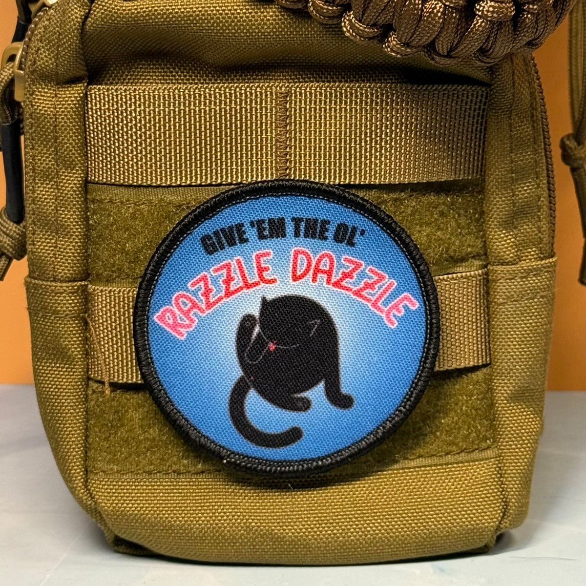 A circular embroidered patch with a black cat illustration and the text 'GIVE 'EM THE OL' RAZZLE DAZZLE' is attached to a tan tactical pouch. The patch has a blue background with red and black text.