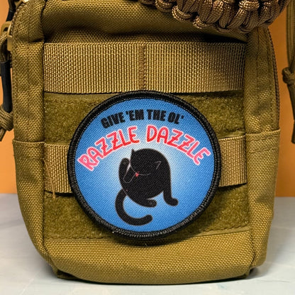 A circular embroidered patch with a black cat illustration and the text 'GIVE 'EM THE OL' RAZZLE DAZZLE' is attached to a tan tactical pouch. The patch has a blue background with red and black text.