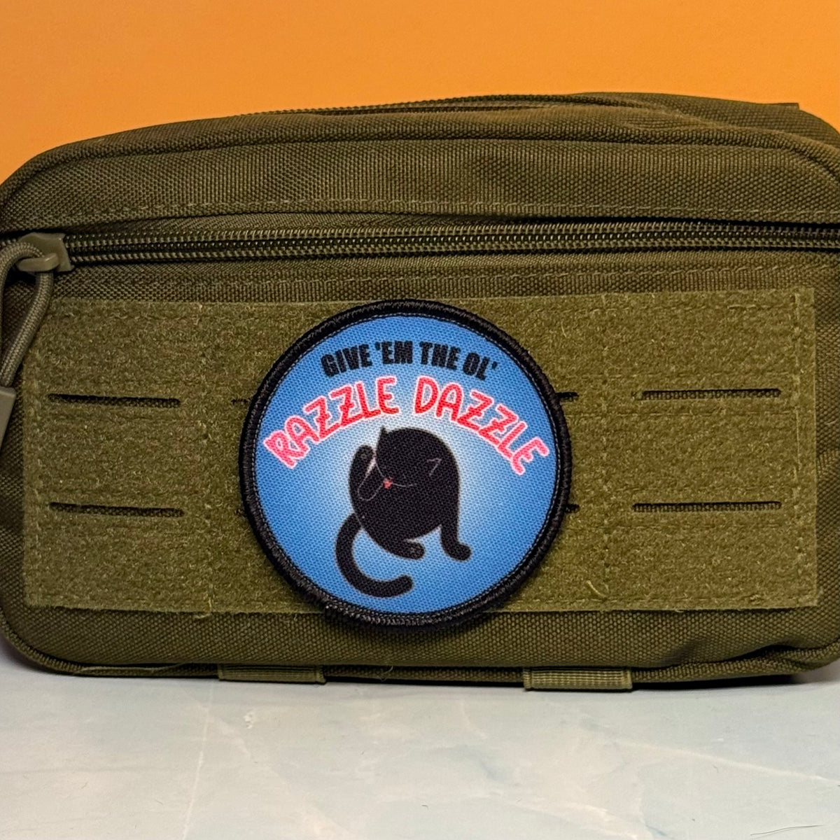 An olive green tactical pouch with a circular blue patch featuring a black cat and the text 'GIVE 'EM THE OL' RAZZLE DAZZLE'.
