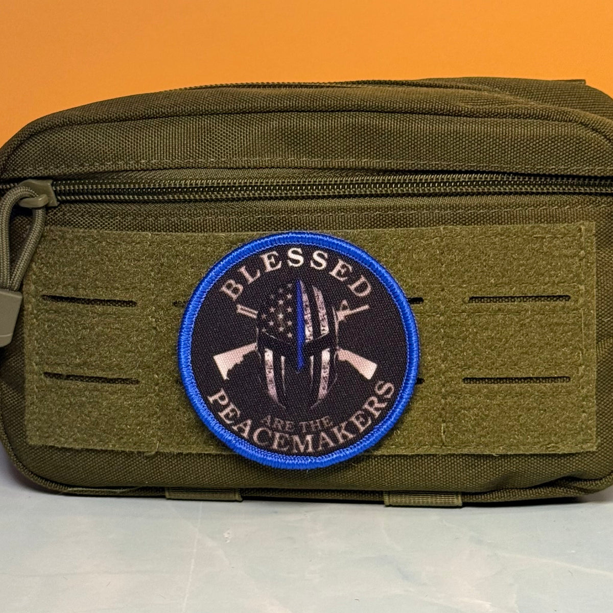 An olive green tactical pouch with a circular embroidered patch reading 'BLESSED ARE THE PEACEMAKERS' in blue, black, and white, set against an orange background.