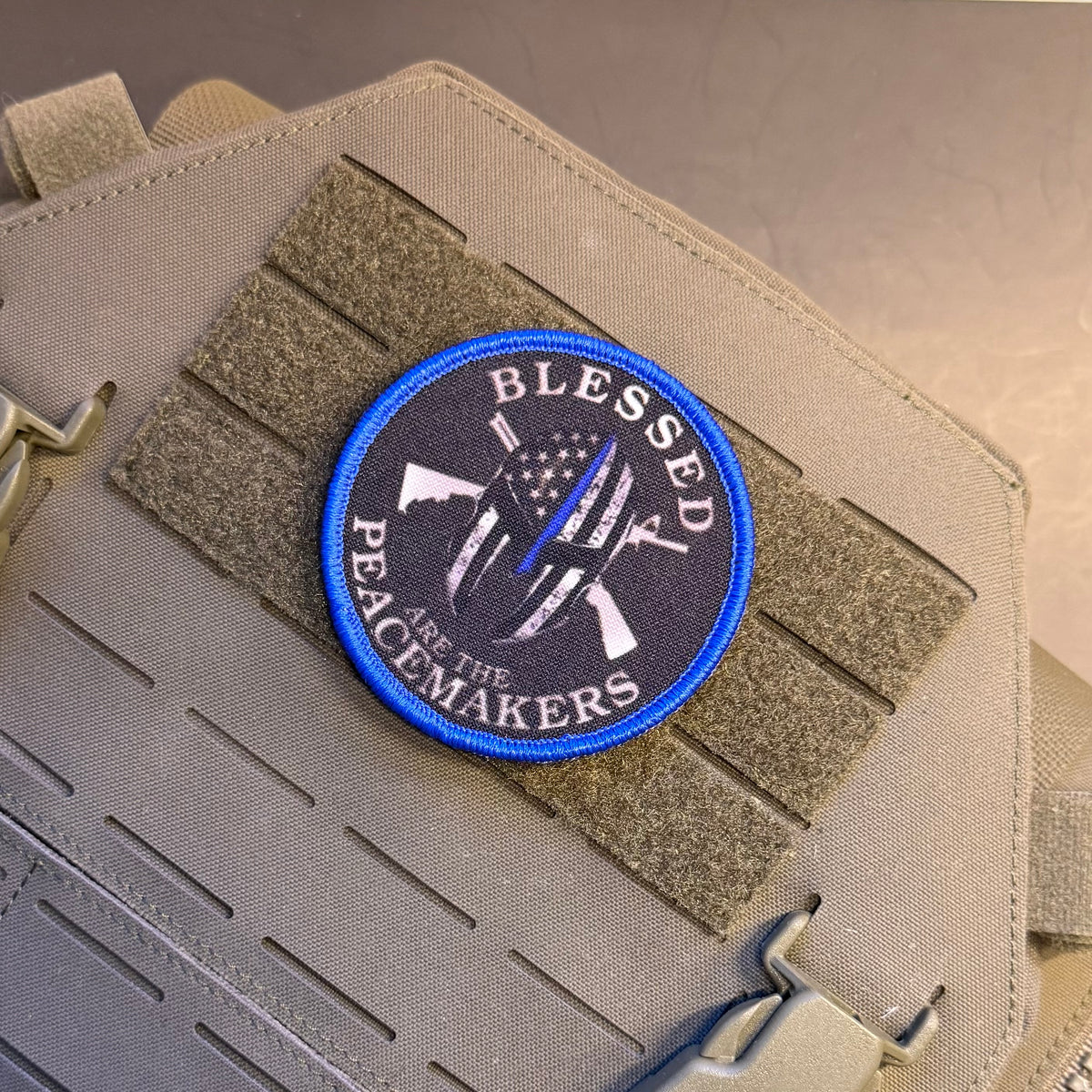 A circular embroidered tactical patch with a blue border and the words 'BLESSED PEACEMAKERS' surrounding a rifle and flag design, shown attached to tan webbing on a beige vest or bag.