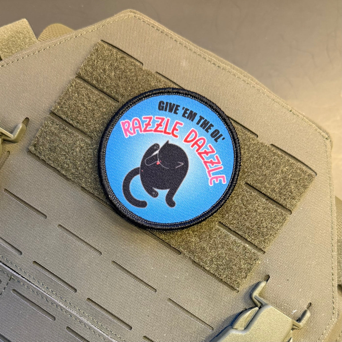 A circular embroidered patch featuring a black cat on a light blue background with the text 'GIVE 'EM THE DO' and 'RAZZLE DAZZLE' in red and black, shown attached to a tan tactical vest with Velcro strips.