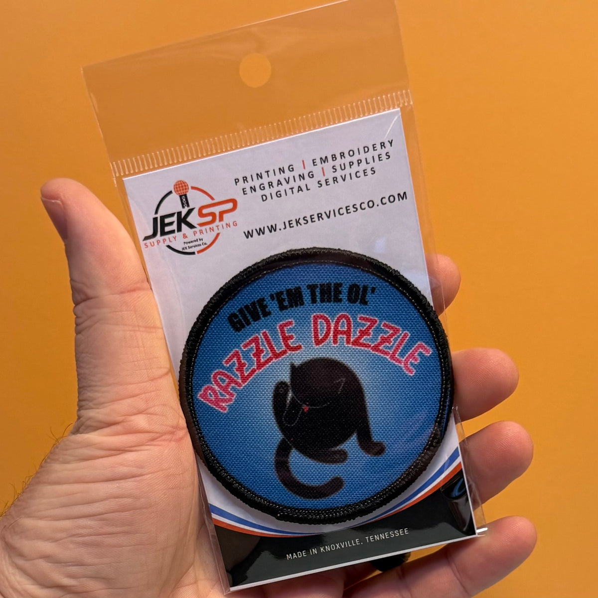 A hand holding a clear plastic blister pack containing a circular embroidered patch. The patch has a blue background with a black cat silhouette and the text “GIVE ’EM THE OL” and “RAZZLE DAZZLE” in red and white.