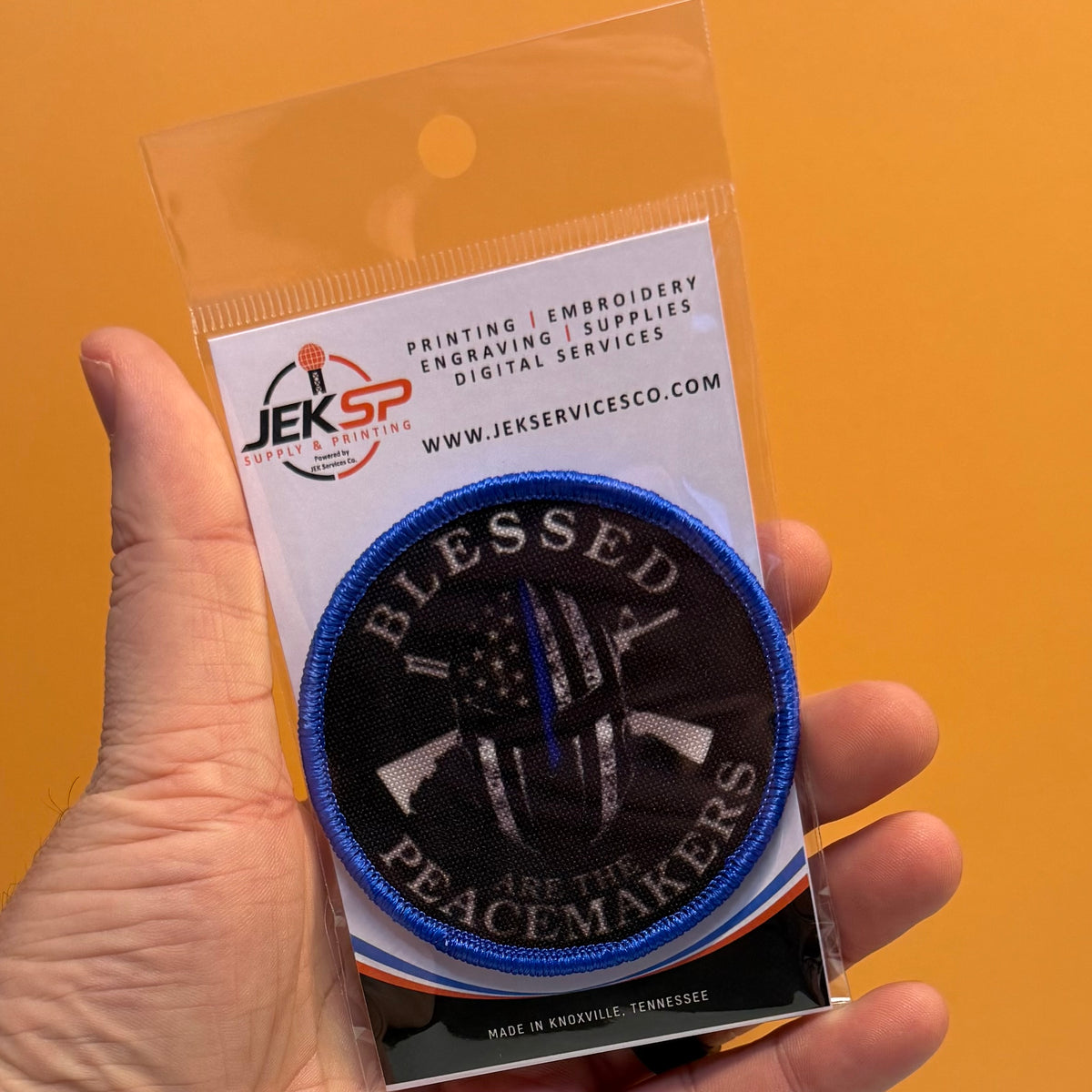 A hand holds a clear plastic pouch containing an embroidered circular patch. The patch reads “BLESSED ARE THE PEACEMAKERS” with a blue border, black background, and a crossed rifle-and-flag design.