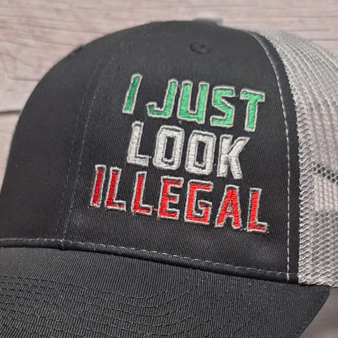 Black cap with 'I Just Look Illegal' text on a wooden surface