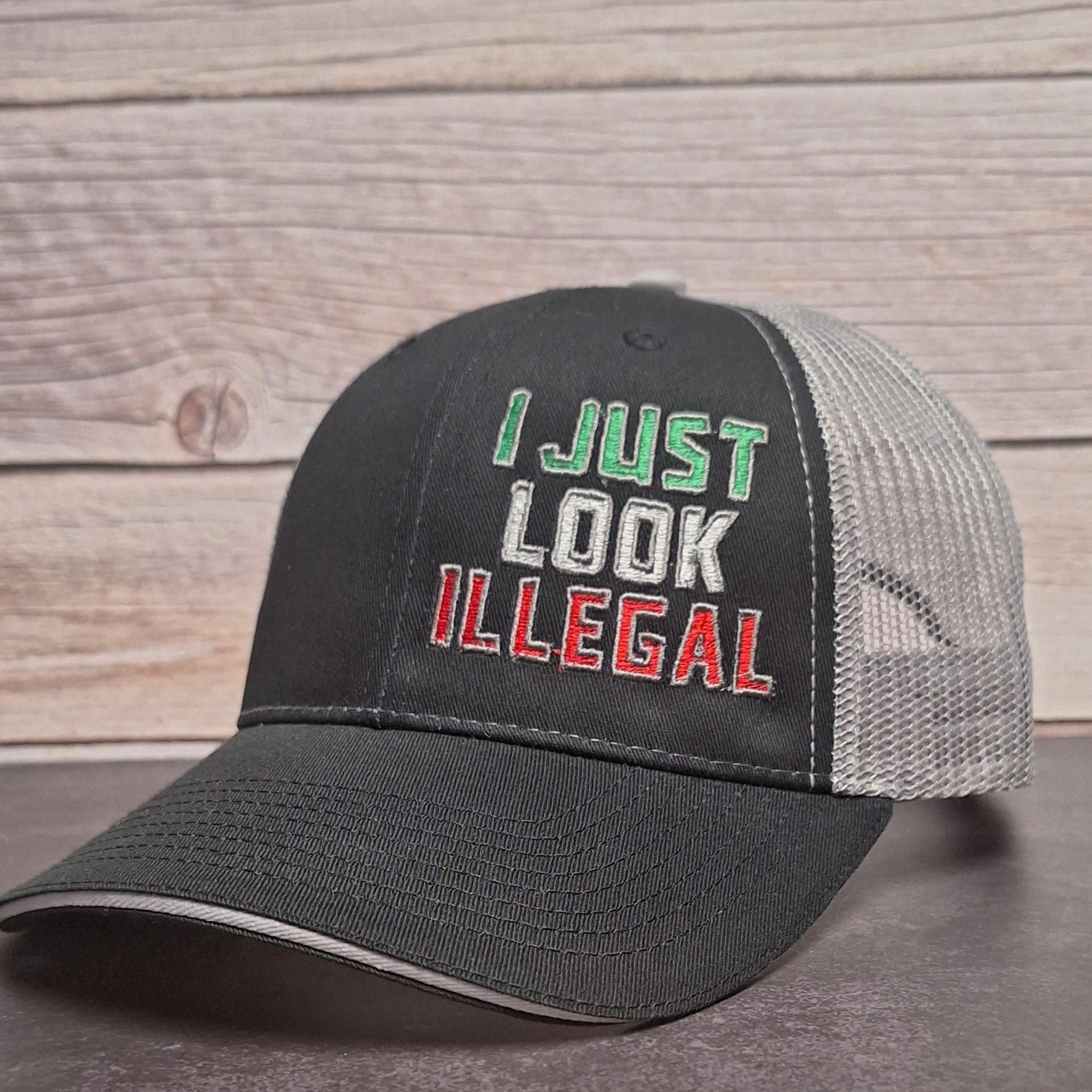 Black and gray cap with 'I Just Look Illegal' text on a wooden surface