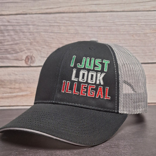 Black and gray cap with 'I Just Look Illegal' text on a wooden surface