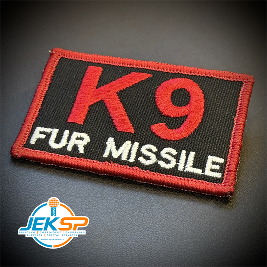 A rectangular embroidered patch with a black background, red border, red 'K9' text, and white 'FUR MISSILE' text, photographed on a dark surface. A JEKSP watermark appears in the lower-left corner.