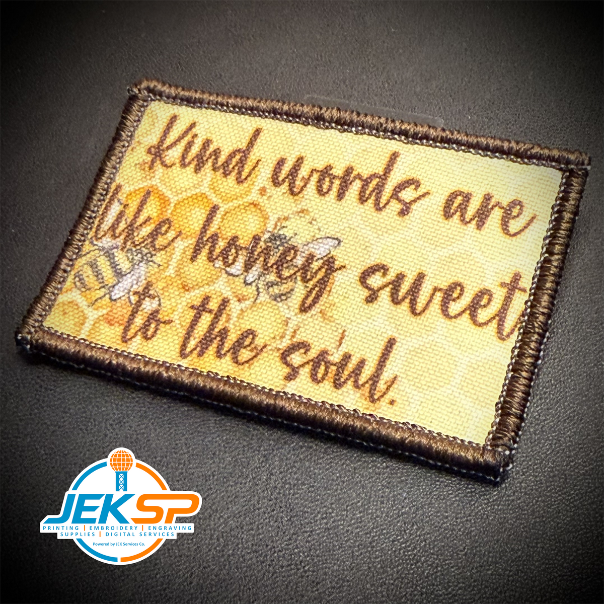 A rectangular embroidered patch with a brown stitched border, featuring the phrase 'Kind words are like honey sweet to the soul.' in brown script over a pale yellow honeycomb and bee design.