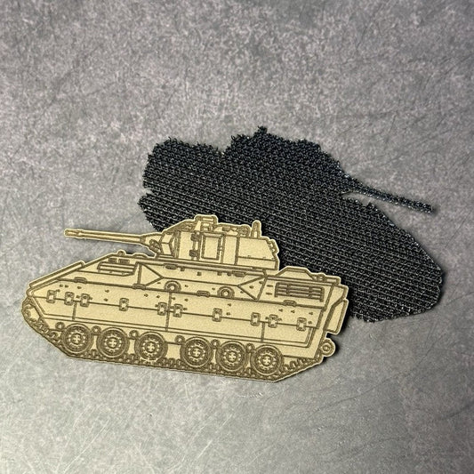 A laser-cut metal enamel pin of a tank vehicle in a light tan color, displayed in front of a black embroidered patch on a textured gray background.