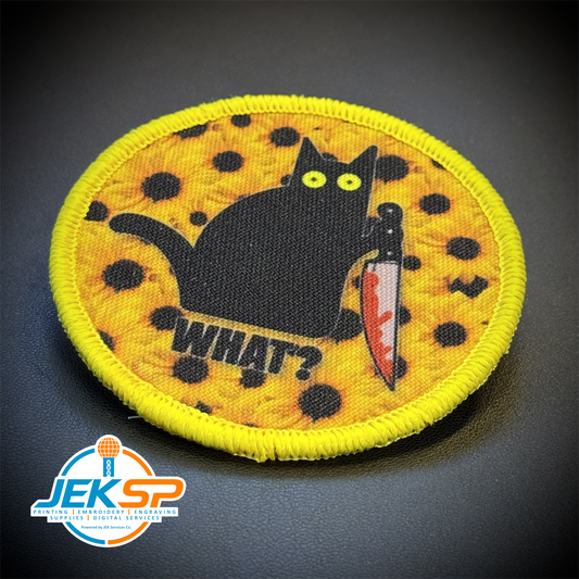 A circular embroidered patch featuring a black cat holding a knife on a yellow spotted background with the word 'WHAT?' below it, photographed on a dark textured surface. A JEKSP watermark is visible in the lower-left corner.