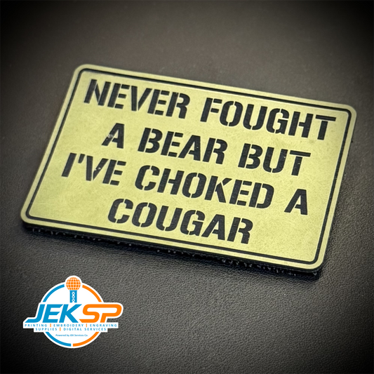 A rectangular enamel lapel pin with a distressed gold background and black text reading 'NEVER FOUGHT A BEAR BUT I'VE CHOKED A COUGAR', displayed on a dark textured surface with a JEKSP brand logo in the bottom-left corner.
