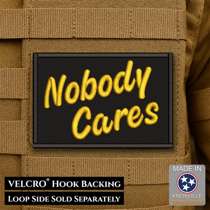 A rectangular black patch with raised yellow embroidered text reading 'Nobody Cares' attached to tan tactical webbing. Text at the bottom states 'VELCRO HOOK BACKING LOOP SIDE SOLD SEPARATELY,' and a 'Made in Knoxville Tennessee' badge appears in the lower right.