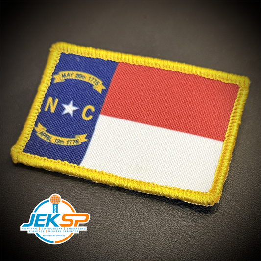 An embroidered rectangular patch featuring the North Carolina state flag with a yellow stitched border, set against a dark textured background. A 'JEKSP' brand watermark is visible in the bottom-left corner.