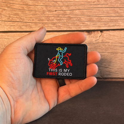 A hand holding a small black embroidered patch with a cowboy riding a bucking bull graphic and the text 'THIS IS MY FIRST RODEO' against a rustic wood plank background.