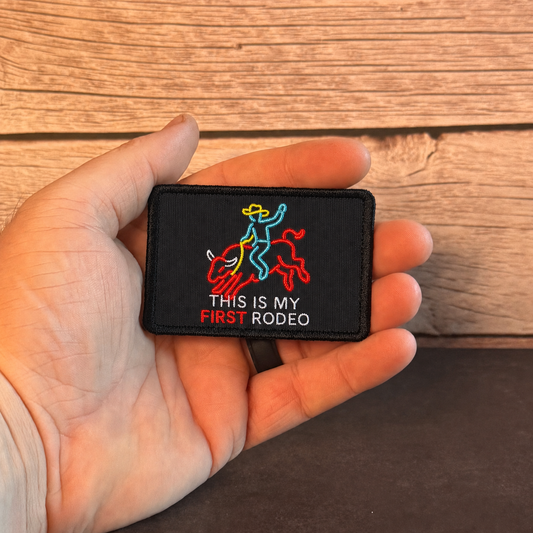 A hand holding a small black embroidered patch with a cowboy riding a bucking bull graphic and the text 'THIS IS MY FIRST RODEO' against a rustic wood plank background.