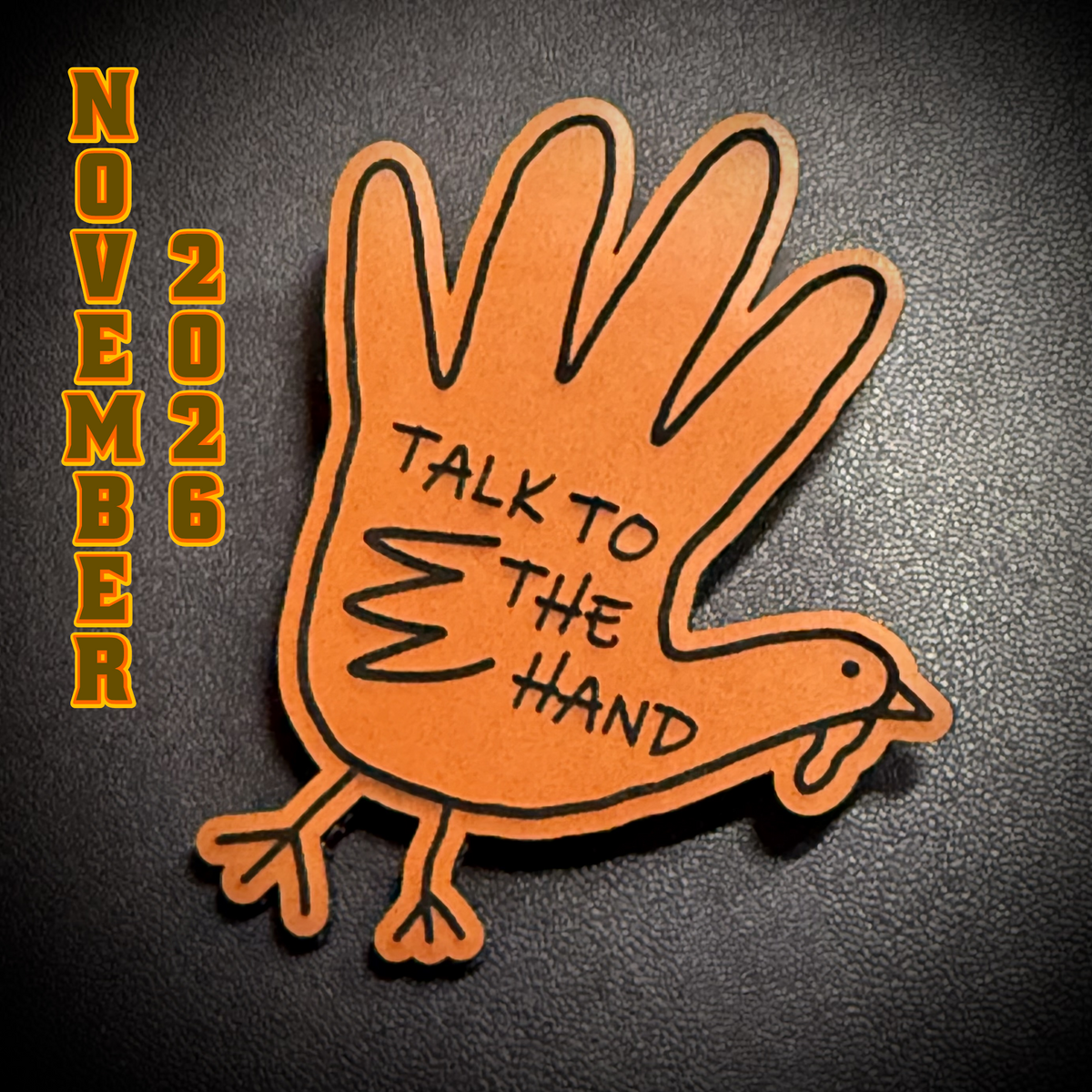 A die-cut paper turkey-shaped sticker or pin with the phrase 'TALK TO THE HAND' written on its body, shown on a dark textured background. Yellow vertical text on the left reads 'NOVEMBER 2026'.