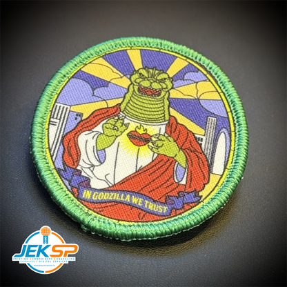 A circular embroidered patch featuring a stylized Godzilla with a green scaly head, red cape, and white robe, set against a yellow and blue sunburst background. The patch has a green stitched border and the text 'IN GODZILLA WE TRUST' at the bottom.