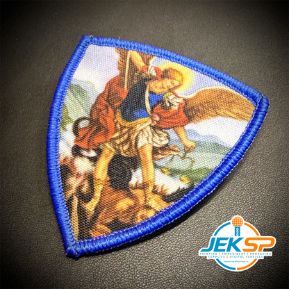 Saint Michael Shield printed canvas morale patch, a triangular printed patch with a blue stitched border and a printed religious scene of a saint or angel in red and blue garments on a dark textured background.
