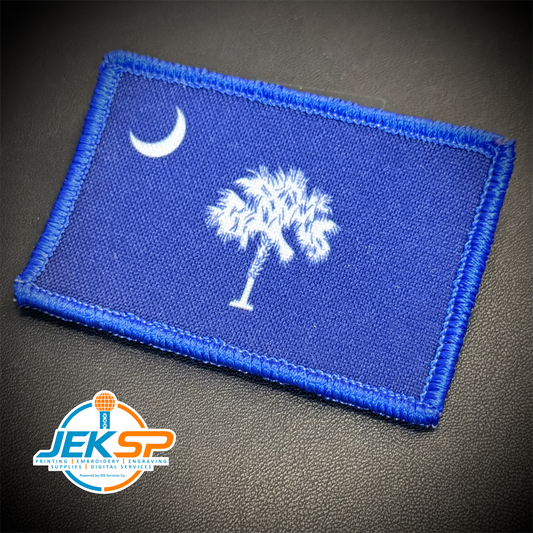 A rectangular embroidered patch featuring the South Carolina state flag design, with a blue field, white crescent moon, and white palmetto tree, photographed on a dark textured surface. A JEKSP watermark is visible in the lower-left corner.