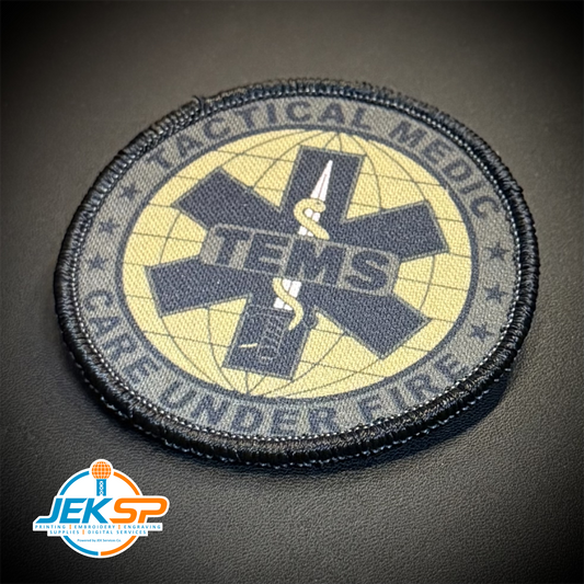 A circular tactical medic patch with a black border and a central black Star of Life symbol over a pale yellow background, featuring the text 'TACTICAL MEDIC' and 'TENS CARE UNDER FIRE.' The patch is photographed on a dark textured surface, with a JEK SP watermark in the lower-left corner.