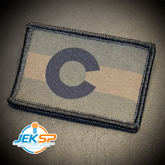 A rectangular tactical patch featuring a dark navy blue letter 'C' over horizontal camouflage stripes in olive green, tan, and dark gray, finished with a black stitched border. The patch is displayed on a dark textured background with a 'JEKSP' brand logo watermark in the bottom-left corner.
