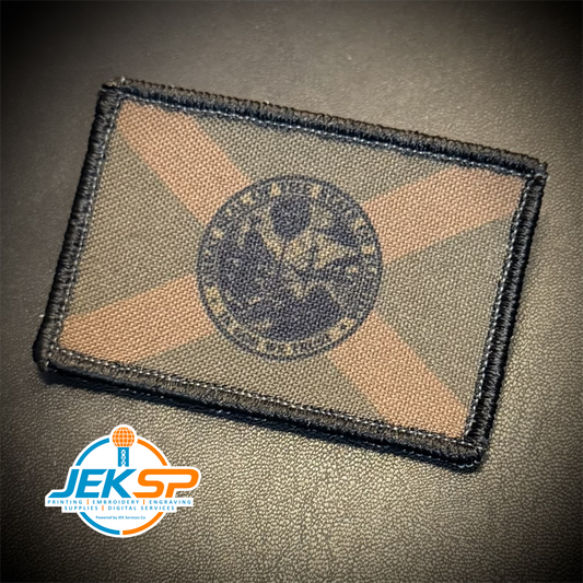 A rectangular embroidered tactical patch featuring a desert camouflage pattern with a black border and a central circular emblem, displayed on a dark textured surface with a JEKSP watermark in the lower-left corner.