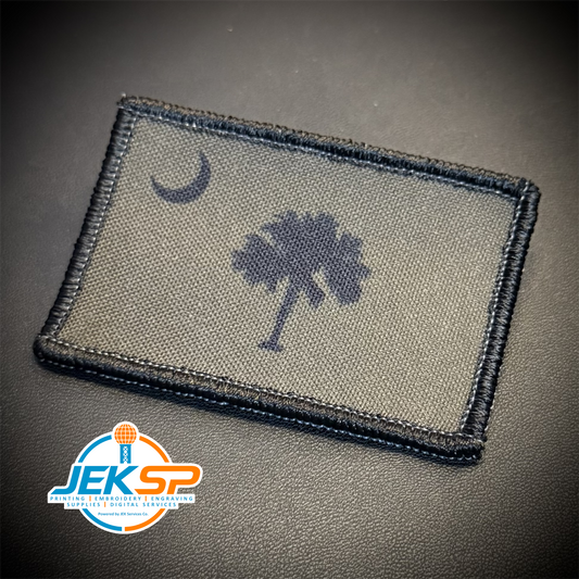 A rectangular tactical patch featuring the South Carolina state flag design with a dark blue crescent moon and palmetto tree on an olive drab background, bordered in black stitching. A 'JEKSP' brand logo is visible in the bottom-left corner.
