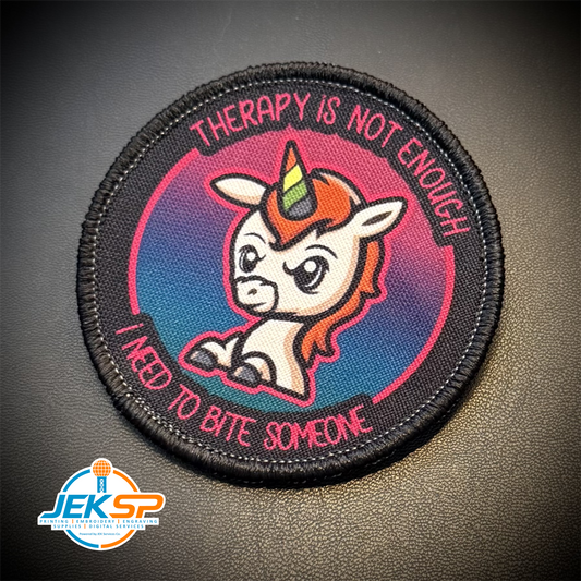 A circular embroidered patch featuring a cartoon unicorn with a rainbow horn, surrounded by the text 'THERAPY IS NOT ENOUGH' and 'I NEED TO BITE SOMEONE' on a black leather-like background. A JEKSP watermark is visible in the bottom-left corner.