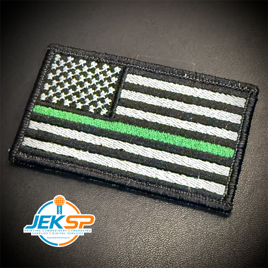 A rectangular embroidered American flag patch with a thin green line, shown on a dark textured surface. The patch has black, white, and green embroidery, with a visible 'JEKSP' logo watermark in the bottom-left corner.
