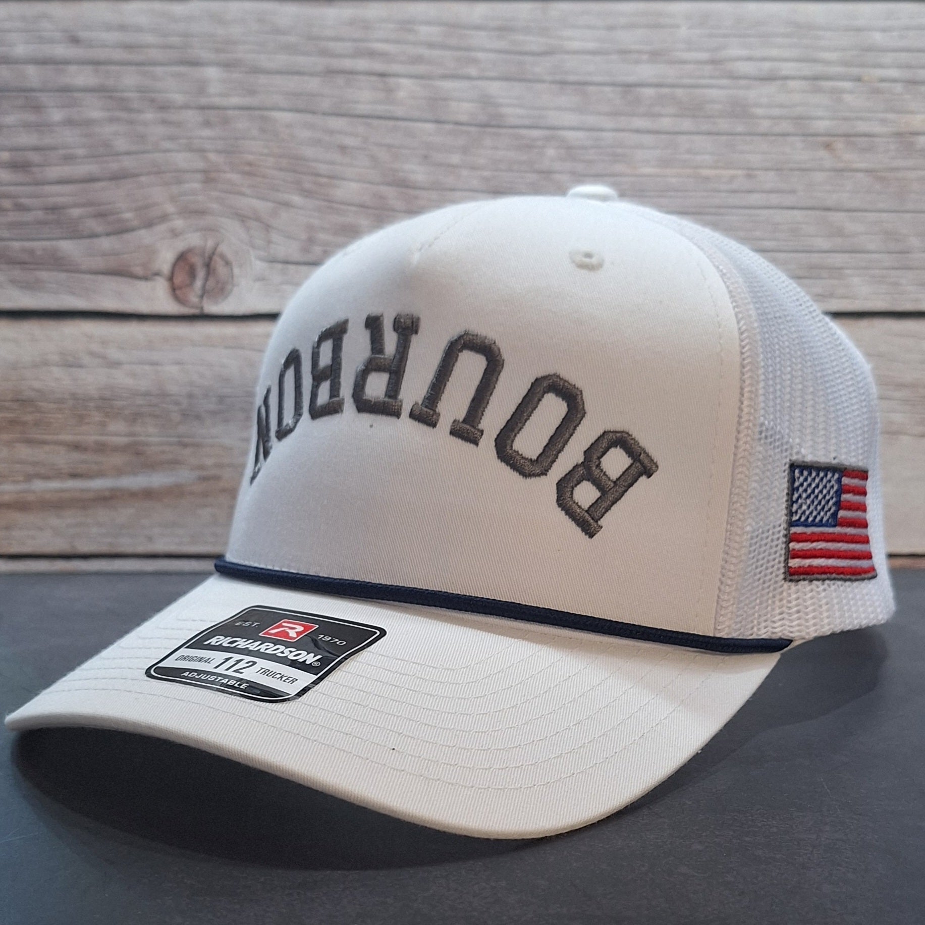 Beige trucker hat with 'Bourbon' text and American flag patch on a wooden surface