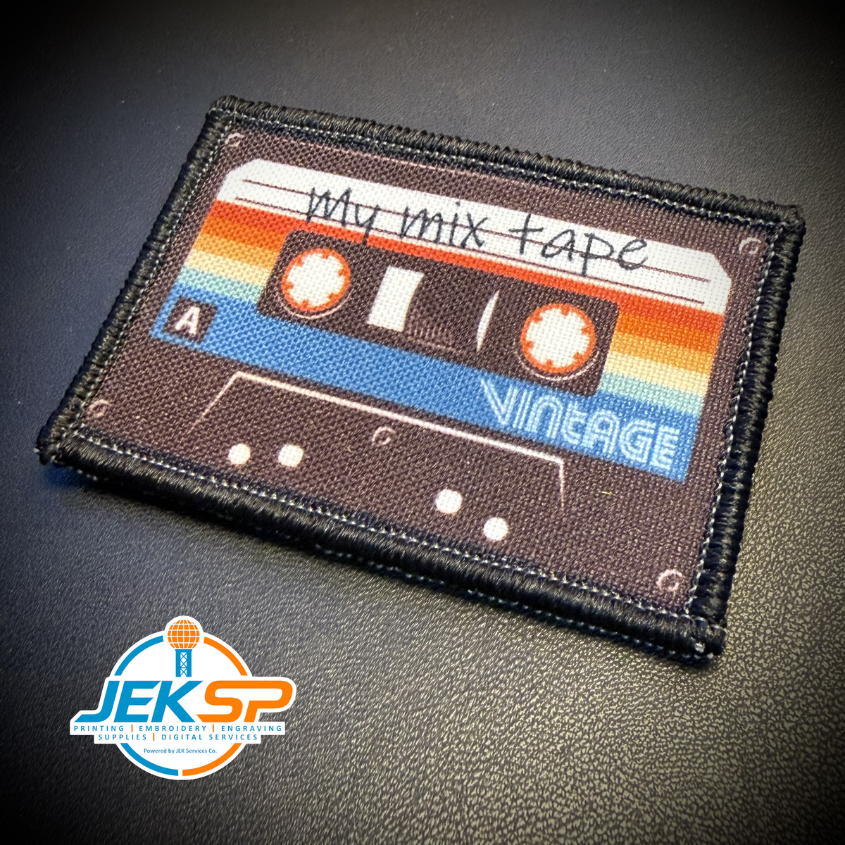 A rectangular embroidered patch shaped like a vintage cassette tape, featuring the text 'My mix tape' and 'VINTAGE' on a dark textured background. The patch is photographed on a black textured surface with a JEKSP watermark in the bottom-left corner.