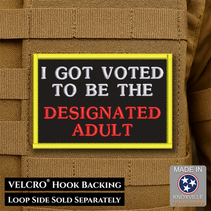 A rectangular embroidered patch with a black center, yellow border, white text reading 'I GOT VOTED TO BE THE', and red text reading 'DESIGNATED ADULT', shown on tan tactical gear. Text at the bottom says 'VELCRO HOOK BACKING LOOP SIDE SOLD SEPARATELY' and a 'MADE IN Knoxville Tennessee' badge appears in the lower right.